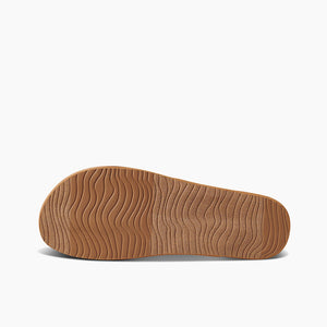 The image shows the bottom view of a REEF Cushion Strand sandal with a light brown sole. The sole features a wavy, ripple-like pattern providing texture and grip. Enhanced arch support and a cushion bounce midsole are highlighted against the plain white background, emphasizing the unique tread design of the shoe sole.
