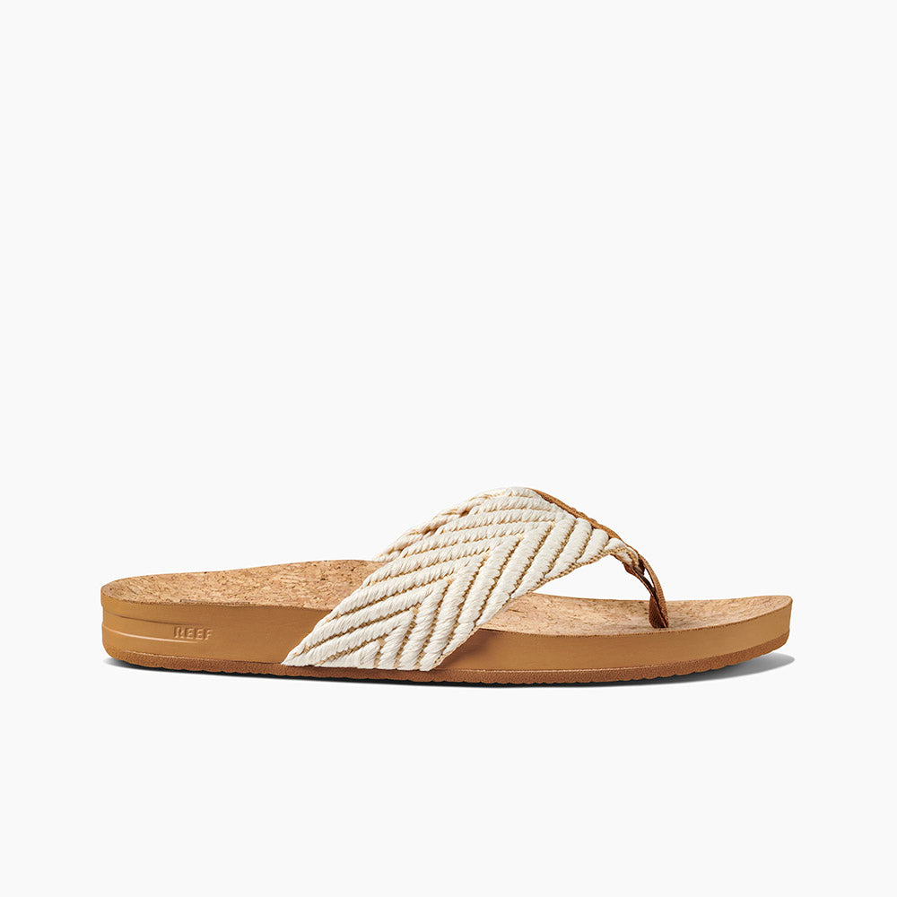 The REEF Cushion Strand is a brown cork-soled sandal with a chevron-patterned cream and white cloth strap. It features a thong design with a brown toe separator, enhanced arch support, and a textured sole for grip. The sandal also includes a cushion bounce midsole for added comfort, with the brand name "REEF" embossed on the side of the sole.
