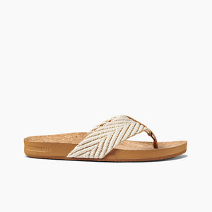 The REEF Cushion Strand is a brown cork-soled sandal with a chevron-patterned cream and white cloth strap. It features a thong design with a brown toe separator, enhanced arch support, and a textured sole for grip. The sandal also includes a cushion bounce midsole for added comfort, with the brand name "REEF" embossed on the side of the sole.