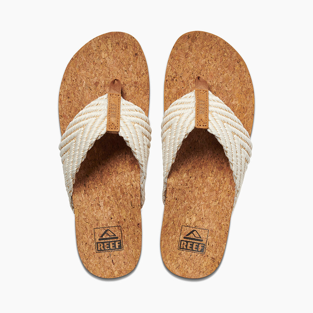 The REEF Cushion Strand sandals feature cork insoles, tan leather accents, and white woven straps. They provide exceptional comfort with their enhanced arch support and cushion bounce midsole. The REEF logo is prominently displayed on the footbeds. The flip-flops are arranged side by side against a white background.