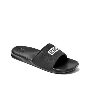 The REEF One Slide is a black sandal featuring a thick strap with a white logo and pebbled surface, showcasing a modern style. Its waterproof design pairs with a textured sole. Positioned at an angle on a white background, it's eco-friendly due to Eco-One biodegradability.