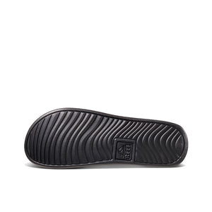 The image features the sole of a REEF One Slide sandal in black, showcasing wavy tread patterns and a prominently displayed brand logo, emphasizing its waterproof design.