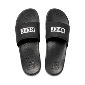 The REEF One Slide sandals are black with "REEF" in white on the straps, featuring Eco-One biodegradability. The textured soles have an embossed logo near the heel. These eco-friendly slides are positioned side by side, facing forward.