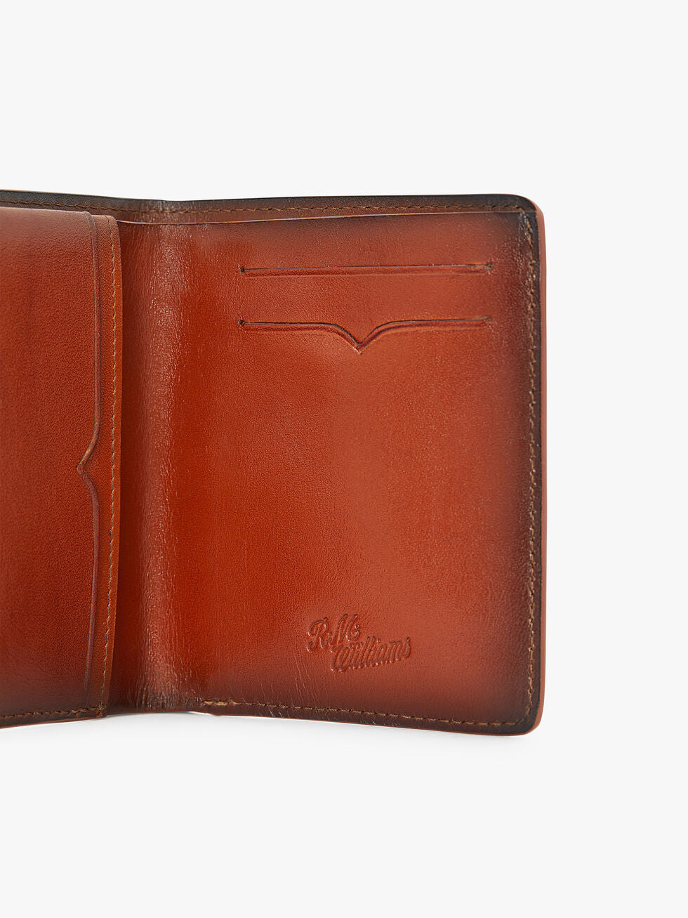 The RM Williams Urban Bi-Fold is a brown full grain leather wallet with two card slots, embossed "R.M. Williams" logo on the lower right, smooth finish, and visible edge stitching.