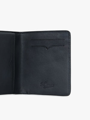 An RM Williams Urban Bi-Fold wallet in black, open to display its full-grain leather interior. The left side features a pocket, while the right side has two card slots. The wallet has embossed writing near the bottom right corner. The background is plain white.