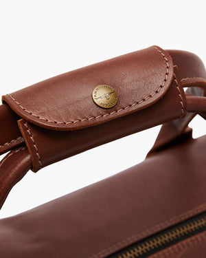 Close-up of the RM Williams Leather Ute Bag's brown leather handle, featuring a stitched flap and round brass "Australia" button. Visible zipper showcases its premium Australian Made craftsmanship.