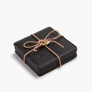 A black RM Williams Heritage 6 Pack Coaster Set is neatly tied with a brown leather cord and adorned with a small brass charm. These Australian-made coasters feature an embossed design, set against a plain white backdrop.
