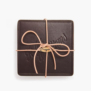 The RM Williams Heritage 6 Pack Coaster Set is a stylish square piece made in Australia, featuring a textured dark leather surface with embossed text. A light tan leather cord, wrapped around the edges and crossed in a loop at the front, enhances its practical design.