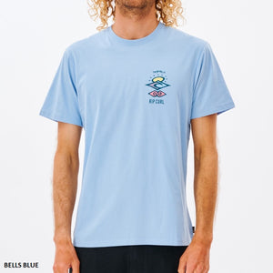 A person with curly blonde hair wears a light blue Rip Curl Search Icon Tee made of organic cotton, displaying a small, colorful graphic design with "Rip Curl" text on the upper left. The photo is cropped from neck to waist against a white backdrop.
