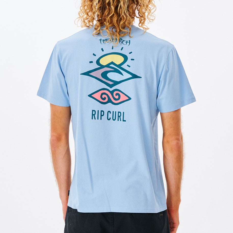 A person with curly hair sports a Rip Curl Search Icon Tee made from organic cotton in light blue, featuring a colorful geometric design and "RIP CURL" text on the back, seen from behind.