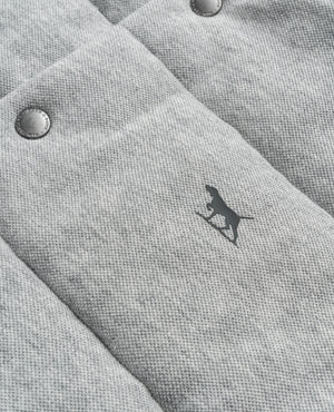 The Rodd & Gunn Jackett Island Vest is a close-up of gray quilted fabric with a dog silhouette. It features two metallic buttons in the upper left corner and an adjustable waist for better fit. The vest offers soft fabric texture with high loft synthetic insulation for warmth and comfort, highlighted by a subtle pattern.