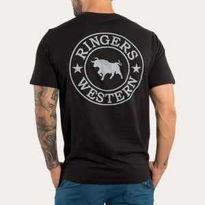 A man with tattooed arms is seen from behind wearing the black Ringers Western Signature Bull Classic T-Shirt, made of 100% cotton, featuring a circular "Ringers Western" logo with a bull and stars. He pairs it with blue pants for a casual look.