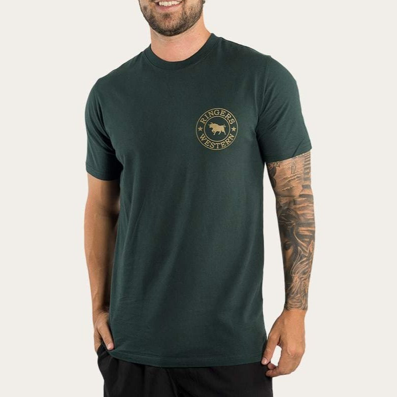A man wears a Ringers Western Signature Bull Classic T-Shirt, dark green with a chest logo, paired with black shorts. A sleeve tattoo marks his right arm. The 100% cotton fabric against the plain white background highlights his relaxed vibe.