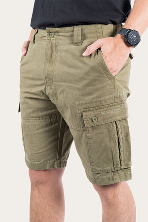 A person is shown from the waist down wearing the Ringers Western Gawler Cargo Short in olive green, made from 100% cotton and featuring multiple pockets. The person has their hands in their pockets and a black watch on their left wrist, with these stylish shorts standing out against the plain light background.