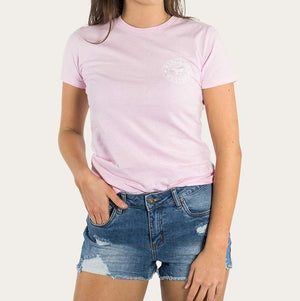 A person is wearing the Ringers Western Womens Signature Bull Classic Fitted T-Shirt in light pink, made from 100% cotton, which features a small circular graphic near the left shoulder. They are also dressed in distressed blue denim shorts, with their right hand resting in the pocket of the shorts and their left hand hanging by their side. The background is plain white.