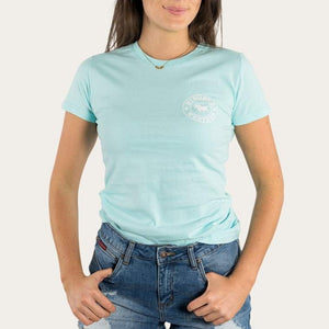 A person wearing a light blue Ringers Western Womens Signature Bull Classic Fitted T-Shirt with a small white circular logo on the left side of the chest and blue jeans, with hands in their pockets. The 100% cotton fabric ensures comfort. The background is plain and light-colored.