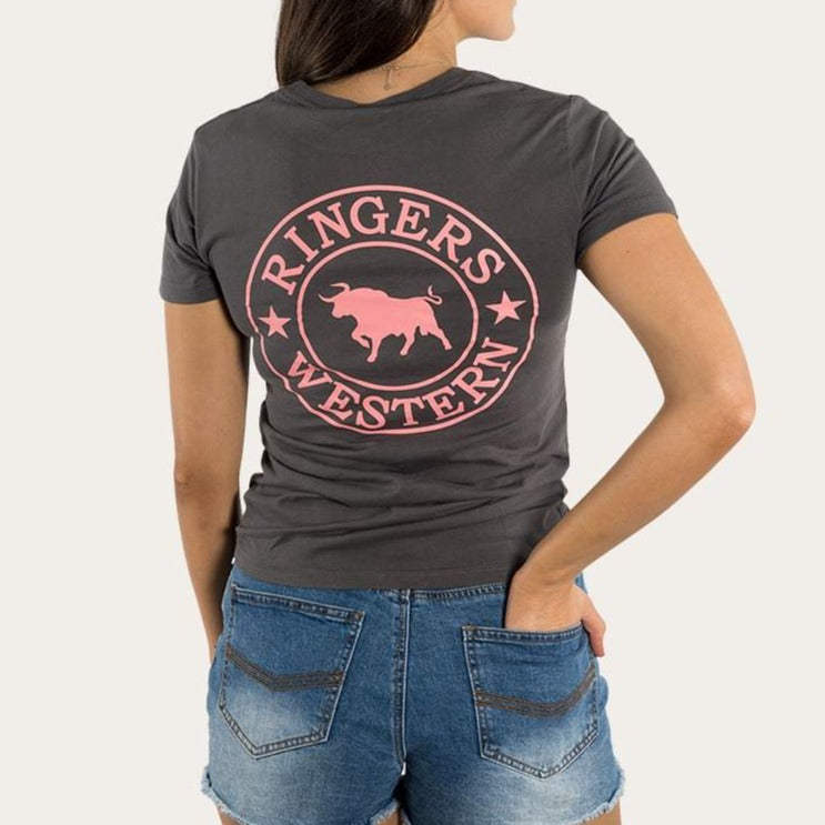A person with long hair stands with their back to the camera, showcasing the "Ringers Western Womens Signature Bull Classic Fitted T-Shirt" in gray, adorned with a pink "Ringers Western" logo featuring a bull. The 100% cotton shirt pairs perfectly with their blue denim shorts, hands resting casually in the pockets.