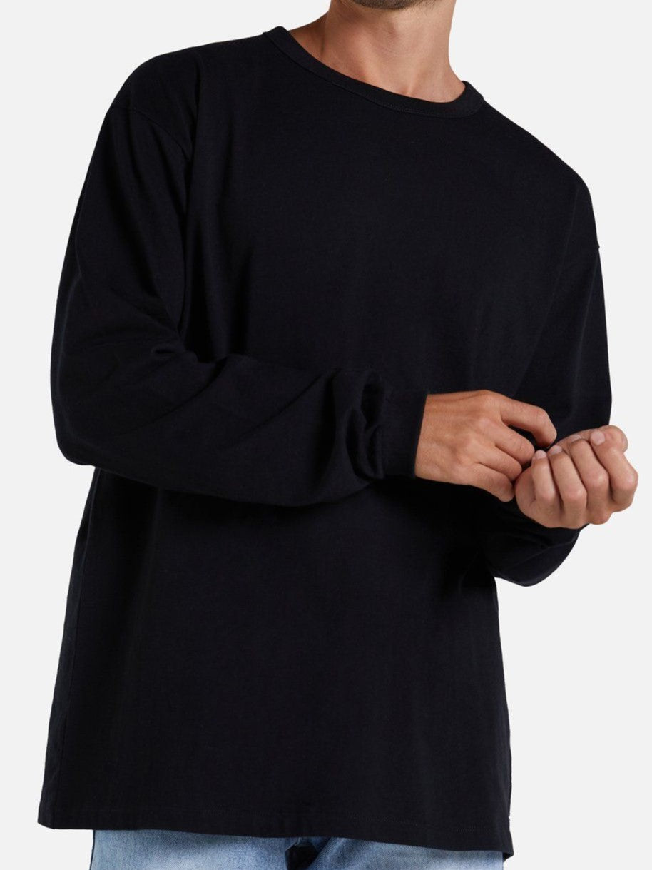A person is shown from the neck down wearing Industrie The Del Sur Long Sleeve Tee in black and light blue jeans. They are adjusting a sleeve of their 100% cotton comfortable tee with one hand against a plain, light-colored background.
