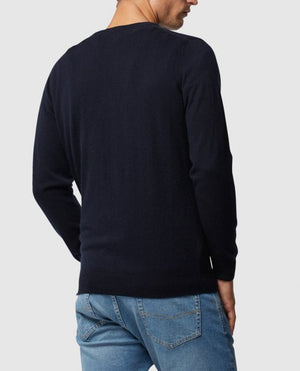 A man wearing a black Rodd & Gunn Queenstown Sweater and blue jeans stands with his back to the camera. The crew neck design enhances the simplicity of the scene against a plain gray background.