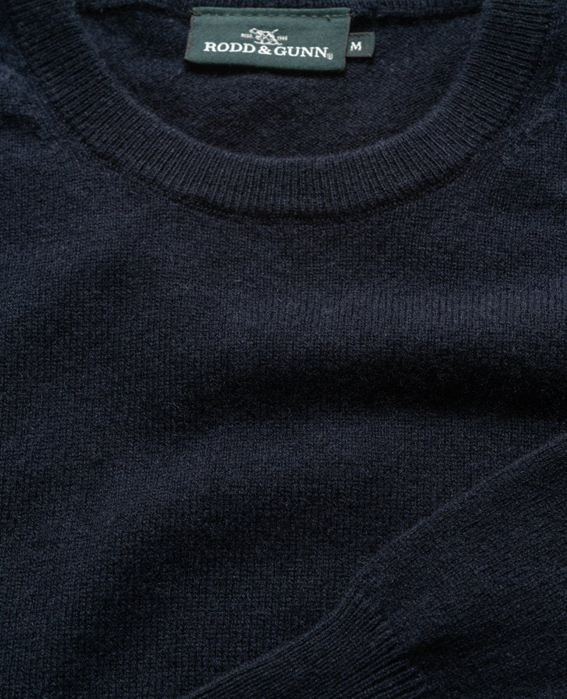 A close-up of a dark blue wool sweater with a crew neck, showcasing the "Rodd & Gunn" tag at the neckline. The knit texture is prominent, and it looks soft, plush, and lightweight; it's the Rodd & Gunn Queenstown Sweater.