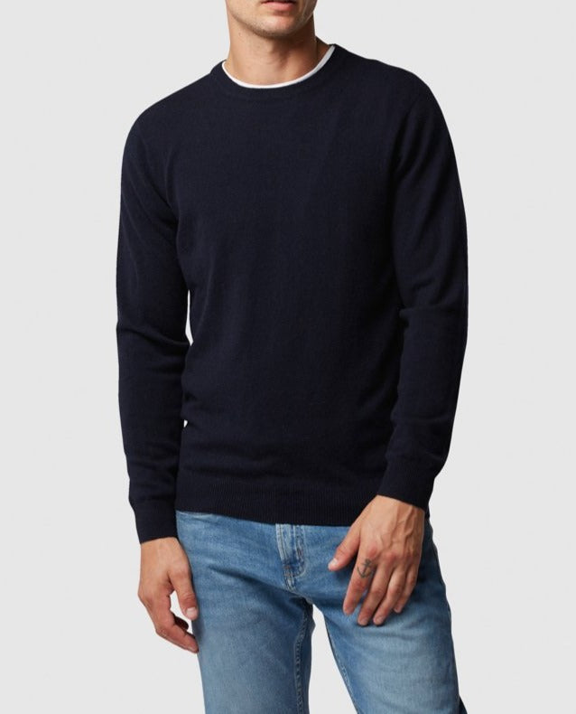 A person in a navy Rodd & Gunn Queenstown Sweater with white-trim crew neck and light blue jeans stands against a white background, left hand slightly tucked into the pocket and right hand resting by their side.