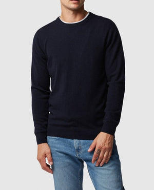 A person in a navy Rodd & Gunn Queenstown Sweater with white-trim crew neck and light blue jeans stands against a white background, left hand slightly tucked into the pocket and right hand resting by their side.