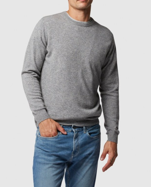 A man wears the Rodd & Gunn Queenstown Sweater in gray wool paired with blue jeans, standing with one hand in his pocket against a plain white background.