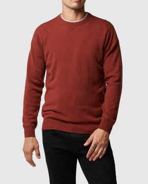 A person stands against a white background in black pants and a rust-colored Rodd & Gunn Queenstown Sweater. The lightweight wool sweater features a round neckline, ideal for layering and travel, as they pose with one hand in front and the other relaxed by their side.