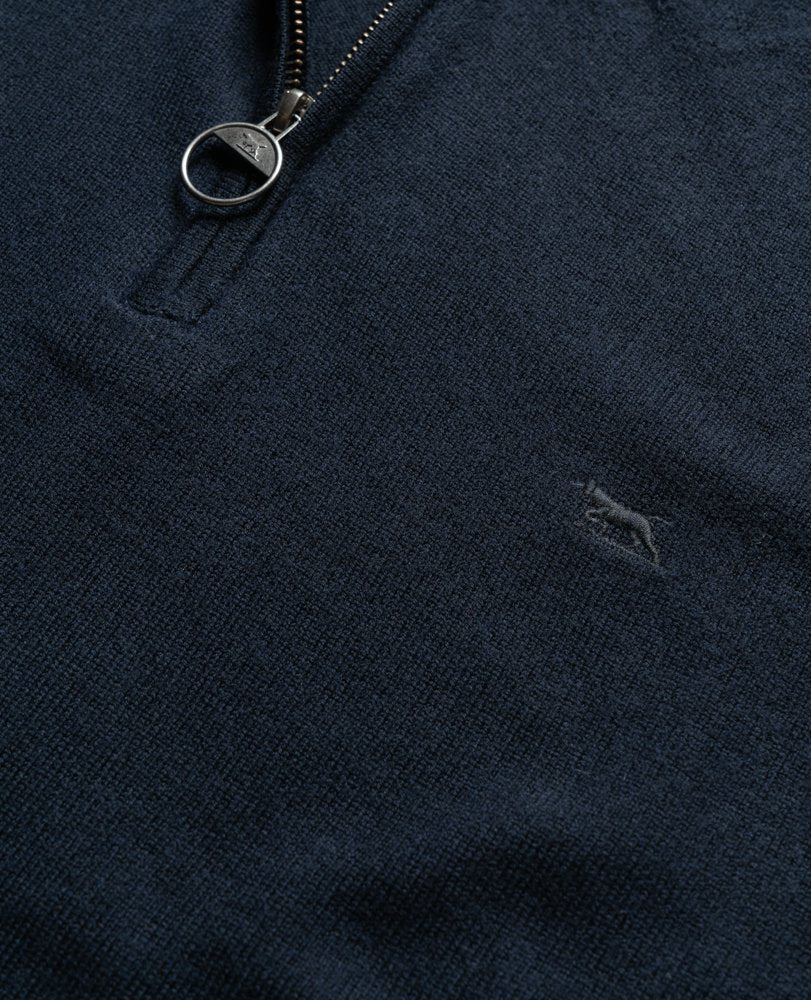 Close-up of a dark blue Rodd & Gunn Bishopsdale Knit with a metal zipper pull ring. The embroidered logo of a small abstract figure in a lighter shade of blue is visible on the lower right section of the Australian Merino quarter-zip knit. The fabric appears to be soft and textured.