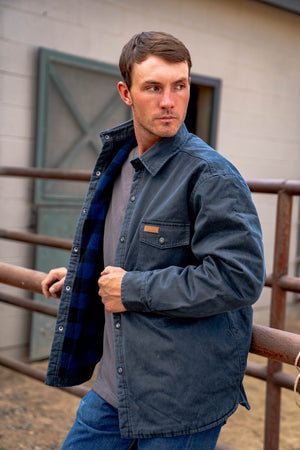 A man leans against a metal fence outdoors, wearing the dark Outback Loxton Jacket with fleece lining over a gray shirt. In the background, a building with a metal door stands. He gazes to the side with short brown hair and a focused expression.