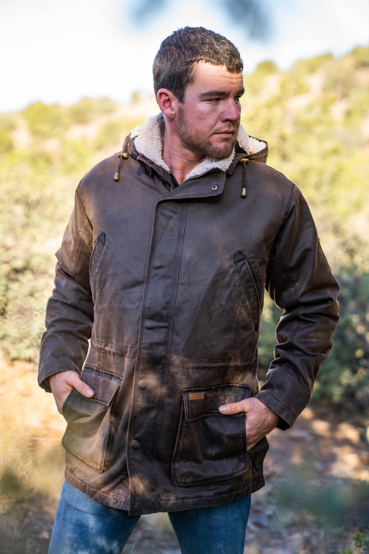A man stands outdoors in the comfortable, water-resistant Outback Nolan Jacket with a furry lining and reinforced elbow patches. He wears blue jeans, looks to the side with hands in pockets, against a sunlit tree backdrop.