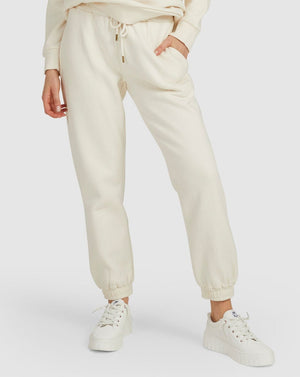 They're wearing Rip Curl Premium Surf Trackpants in cream with an elastic waist, matching hoodie, and partially pocketed hands. White sneakers with thick soles complete their look against a plain light gray background, perfectly capturing the relaxed vibe.