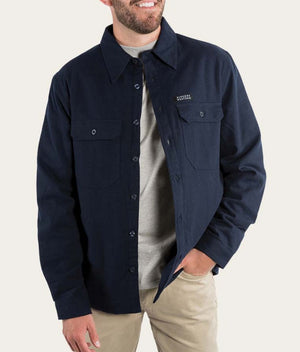A person wearing a Ringers Western Skipton Lumber Jacket in navy blue over a light grey shirt and beige pants. The casual jacket has a collar and two chest pockets with buttons, and the person has their right hand in their pants pocket.