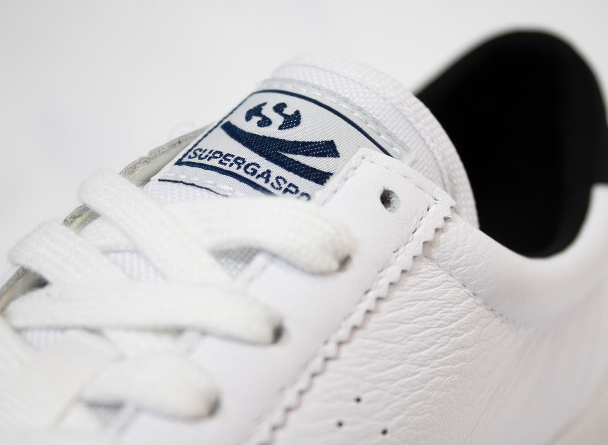 Close-up of a sleek white leather sneaker with pristine laces, showcasing a stitched label on the tongue featuring Superga's circular logo and "SUPERGASPO." The fine texture and intricate stitching highlight the Superga Clubs Comfleasueu Sneaker.
