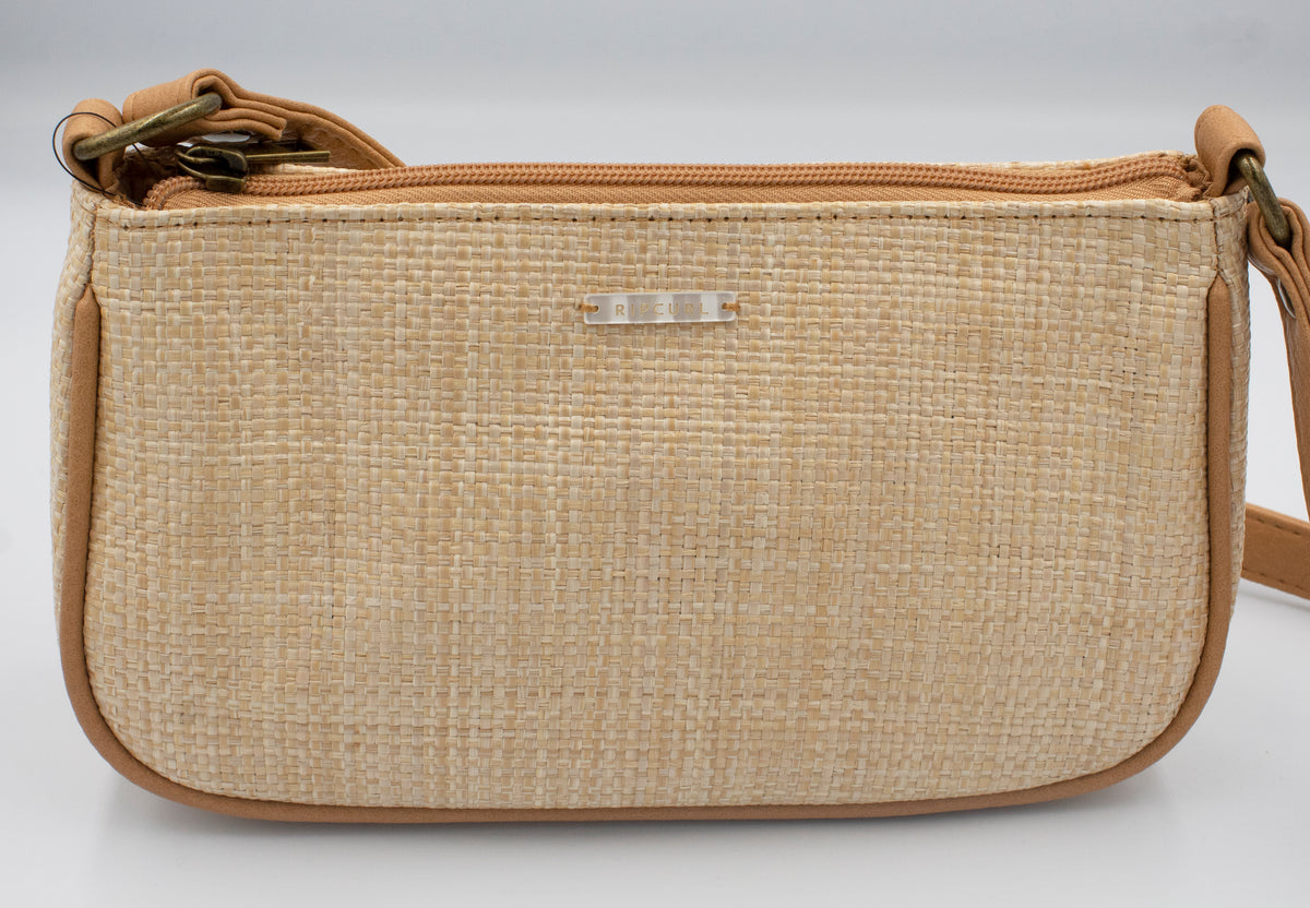 The Rip Curl Sunset Palms Shoulder Bag features a beige woven beachy design with tan leather trim, a shoulder strap, and a small metallic logo on the front. It has a zipper closure and is displayed against a plain white background.