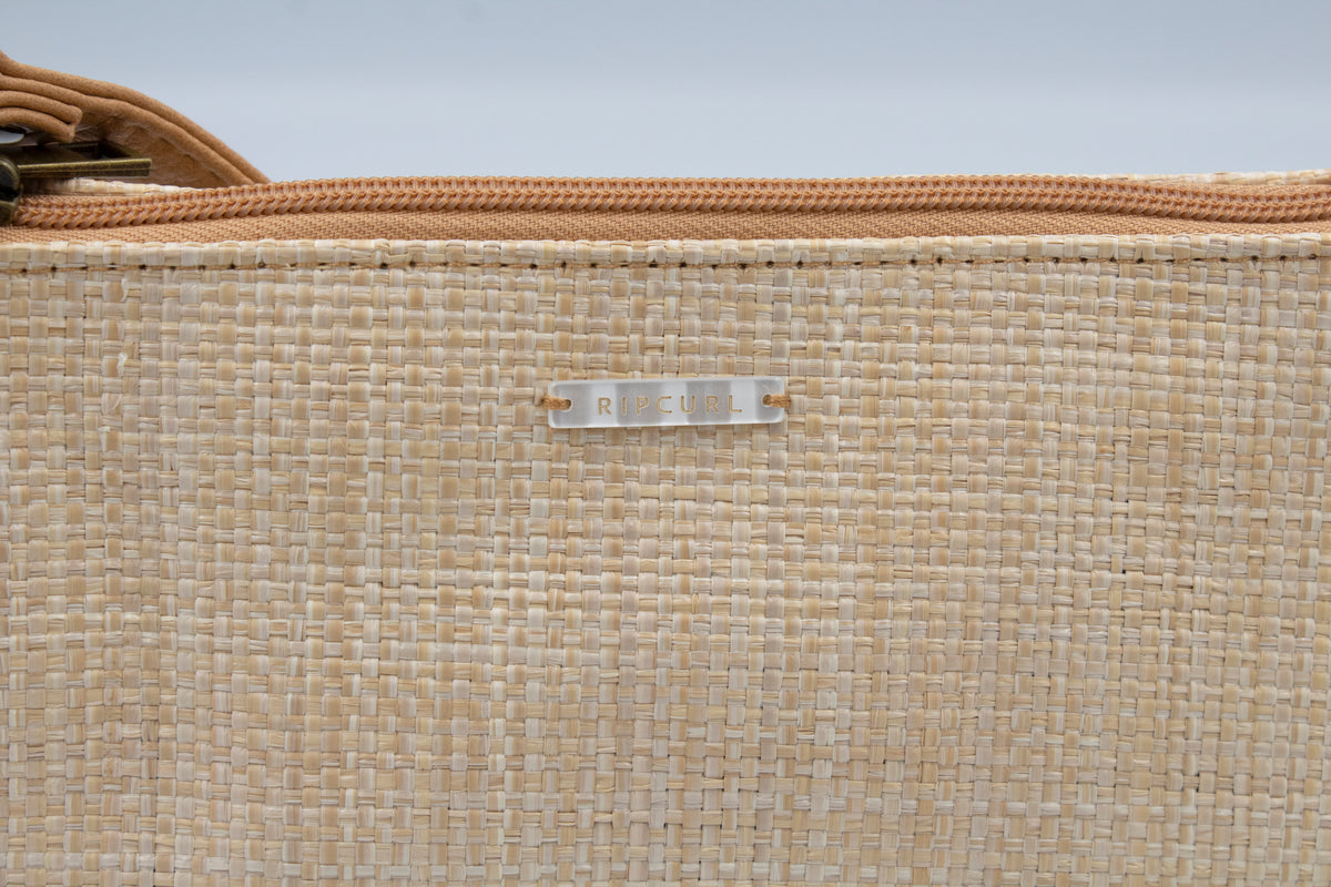 Close-up of the beachy beige woven texture of the Rip Curl Sunset Palms Shoulder Bag, featuring a central silver label saying "Rip Curl." A light brown zipper adorns the top edge, suggesting elegance and function.