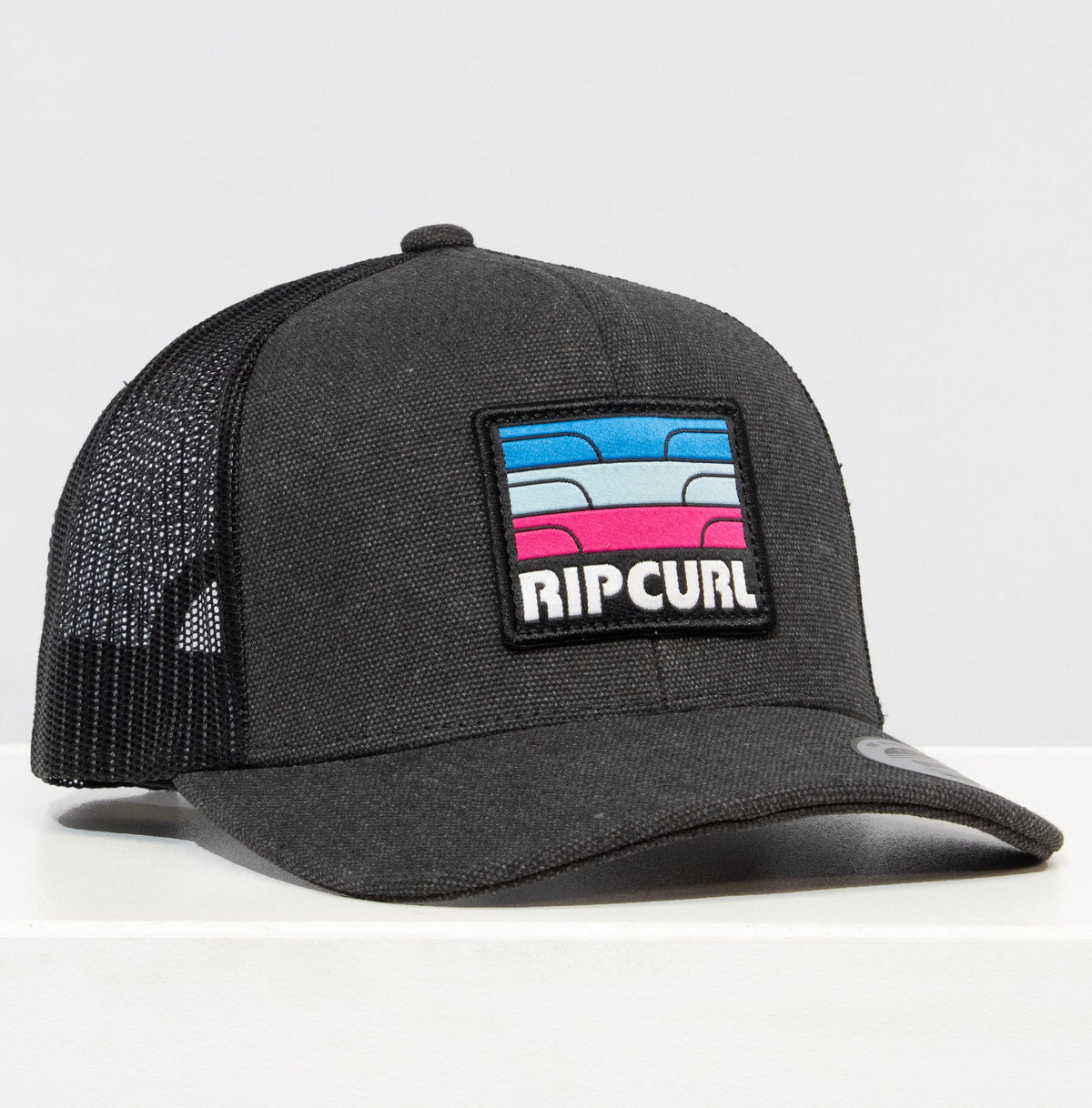 The Rip Curl Surf Revival Curve Trucker hat features a black mesh design adorned with a colorful patch of abstract waves in blue, pink, and red, highlighted by the bold "RIP CURL" logo. It is displayed on a white surface against a plain backdrop.