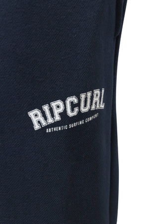 Close-up of Rip Curl Varsity Pants, featuring dark blue fabric with "Rip Curl Authentic Surfing Company" in white text. Ensure the perfect fit by selecting the correct size and consult customer service for any sizing inquiries.
