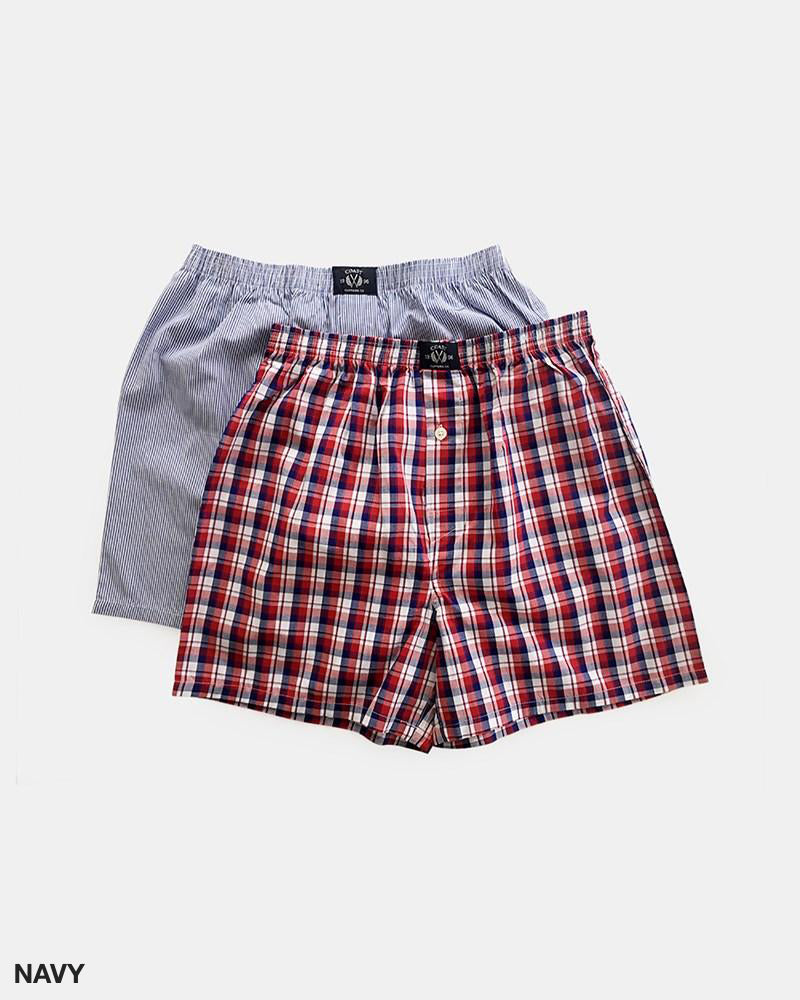 Two pairs of men's boxers from Coast Clothing's Woven Check Boxers 2 Pack collection. One pair is navy blue and white with thin vertical stripes, while the other sports a red, white, and blue woven check pattern. Both 100% cotton boxers are equipped with an elastic waistband and a front buttoned fly. The background is plain white.