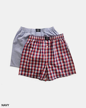 Two pairs of men's boxers from Coast Clothing's Woven Check Boxers 2 Pack collection. One pair is navy blue and white with thin vertical stripes, while the other sports a red, white, and blue woven check pattern. Both 100% cotton boxers are equipped with an elastic waistband and a front buttoned fly. The background is plain white.