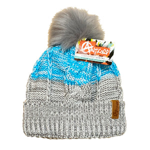 The Anticorp Ladies Freshies Beanie showcases intricate blue, white, and gray patterns, topped with a large, fluffy gray pom-pom. It features a tag with the "Anticorp" brand logo and details. The beanie is complete with fleece lining and a folded brim adorned by a small leather patch on the front.