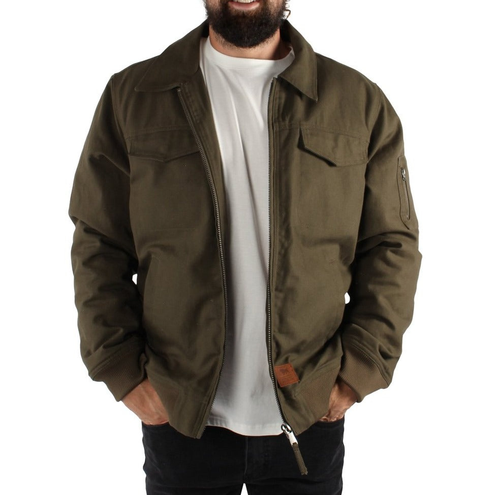 A bearded man is smiling while standing with his hands in the pockets of a military green Ringers Western Mildura Sherpa Bomber Jacket, which features a Durable Water Resistant finish and sherpa lining. He is also wearing a white t-shirt and black pants. The jacket has a zipper and two chest pockets with flaps.