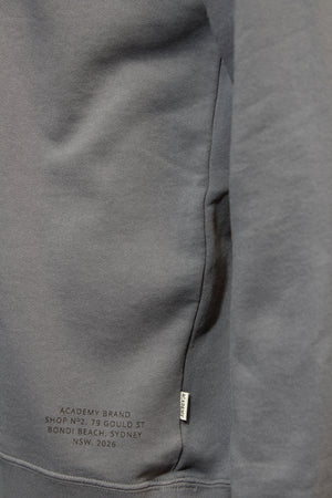 Close-up of a grey Academy Brand Bondi 1/2 Zip Sweat showing a small tag on the seam and printed text: "ACADEMY BRAND, SHOP 1, NO. 79 GOULD ST, BONDI BEACH, SYDNEY, NSW 2026.