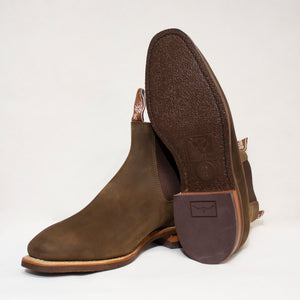 Two brown RM Williams Comfort Craftsman Boots in Crazy Horse leather; one stands upright while the other leans, displaying a textured rubber sole with logo and size mark. The background is plain white.