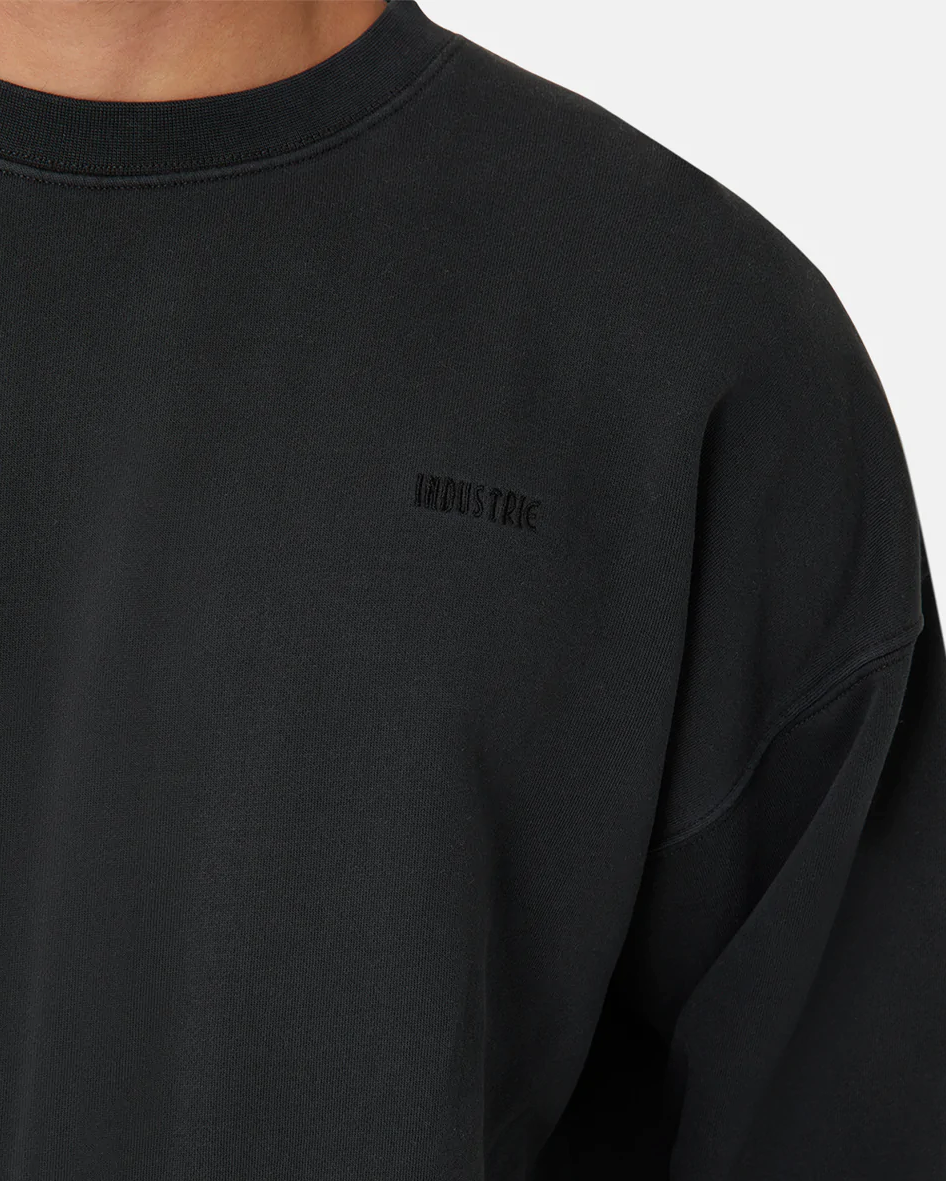 Close-up of a person wearing Industrie The Del Sur Sweat, an oversized black sweatshirt with an acid wash finish. It has subtle "INDUSTRIE" text on the left chest. The plain white background highlights the garment's unique texture.
