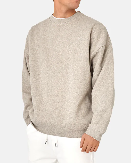 Someone is wearing the Industrie The Del Sur Sweat in light gray with long sleeves and subtle chest embroidery, paired with white pants. The oversized fit and plain light gray background enhance the outfit's relaxed vibe.