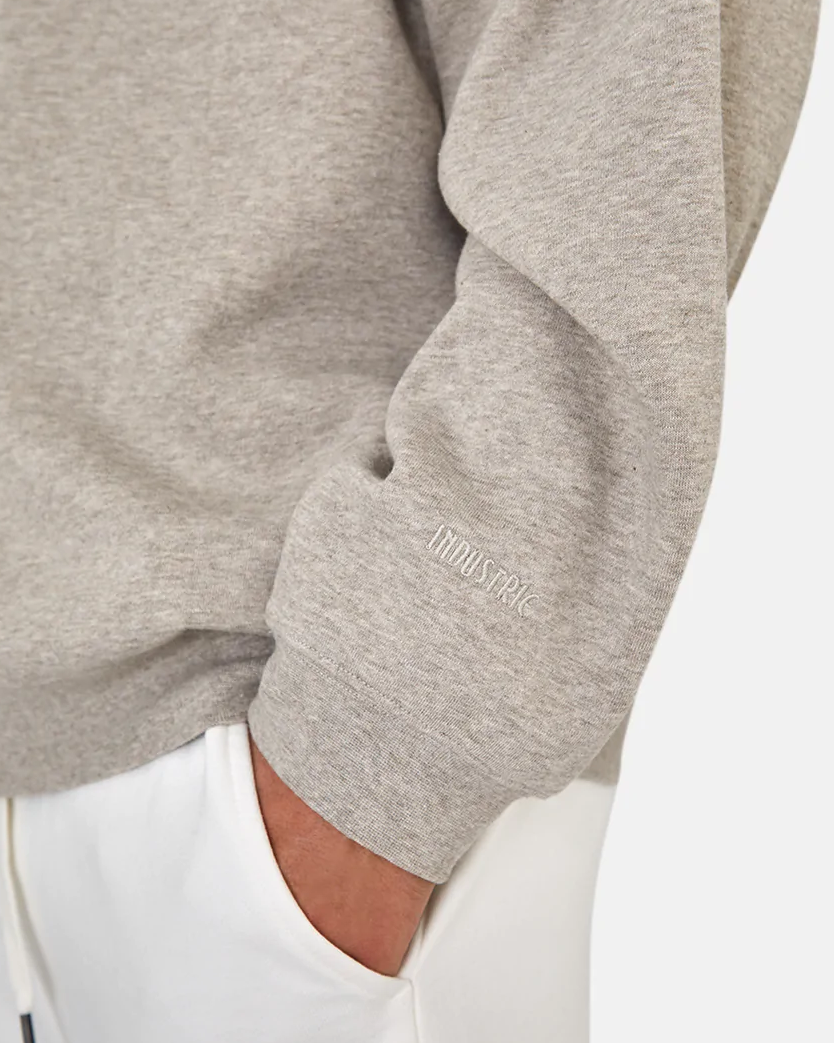 In a close-up, a person dons the oversized Industrie The Del Sur Sweat in light gray with "INDUSTRIE" embroidered on the sleeve, paired effortlessly with white pants. Their arm bent and hand resting by the waist highlight a comfortable and laid-back style.