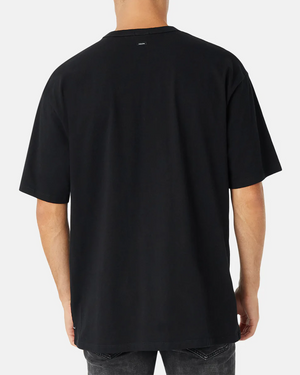 A rear view shows a person in the Industrie The Del Sur Tee, a plain black T-shirt made from 100% cotton, paired with dark jeans against a light gray background. It features a small logo at the top center near the neckline.