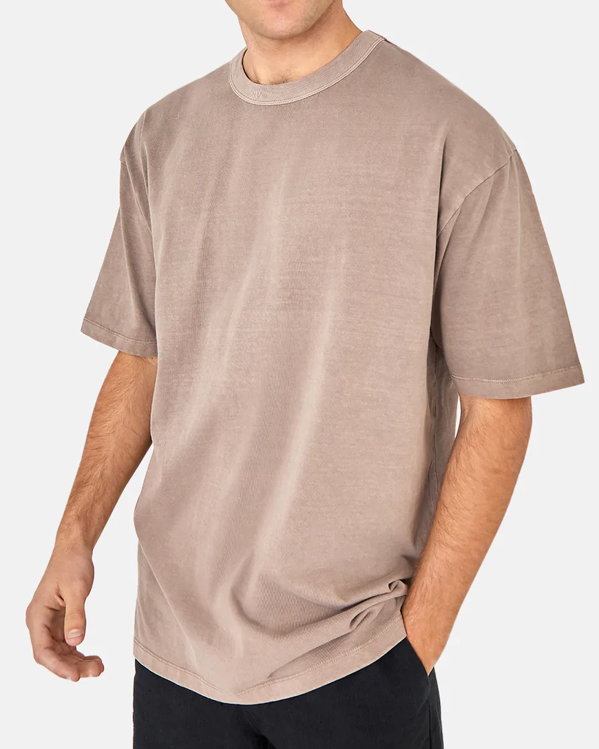 A person models the Industrie The Del Sur Tee, an oversized fit, short-sleeved beige tee made from 100% cotton, paired with black pants. In a cropped photo against a plain background, one hand rests in their pocket while the face remains unseen.