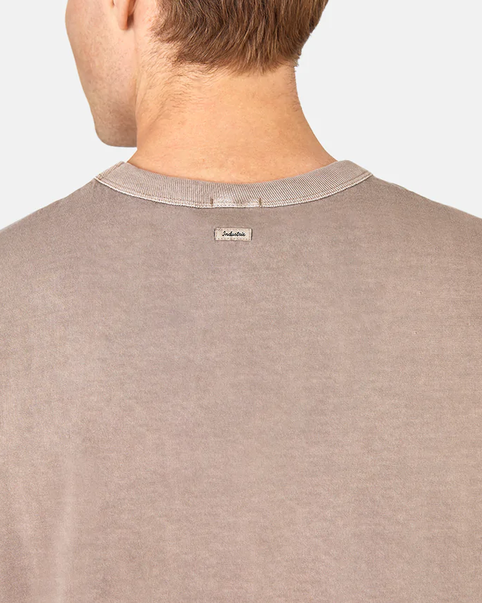 A person with short light brown hair is seen from behind wearing the taupe Industrie The Del Sur Tee with an oversized fit. A small rectangular tag is on the back of the collar, set against a plain light gray background.